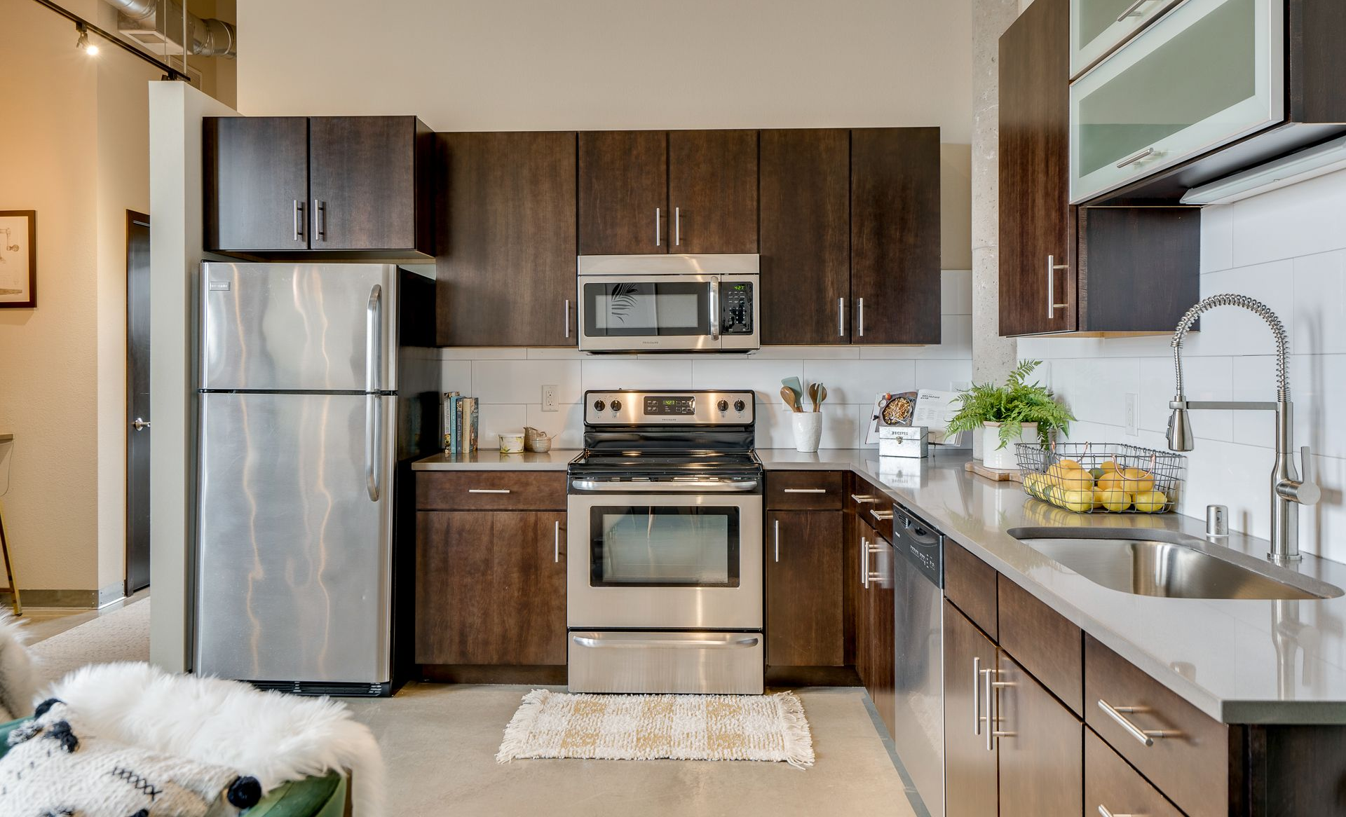 A modern kitchen with dark wood cabinets, stainless steel appliances, and gray countertops, featuring a sink and fruit.