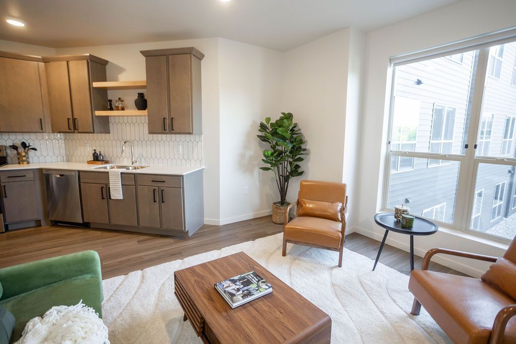 A modern living space with a kitchen featuring brown cabinets, a green sofa, two leather chairs, and a wooden coffee table.