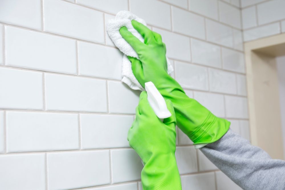 Person wearing green gloves cleaning white tiled wall with a white cloth and spray bottle.