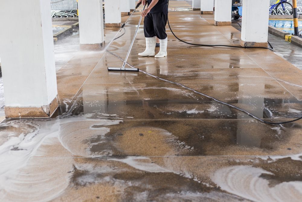 Person in black clothing and white boots cleaning a wet, tiled floor with a squeegee.