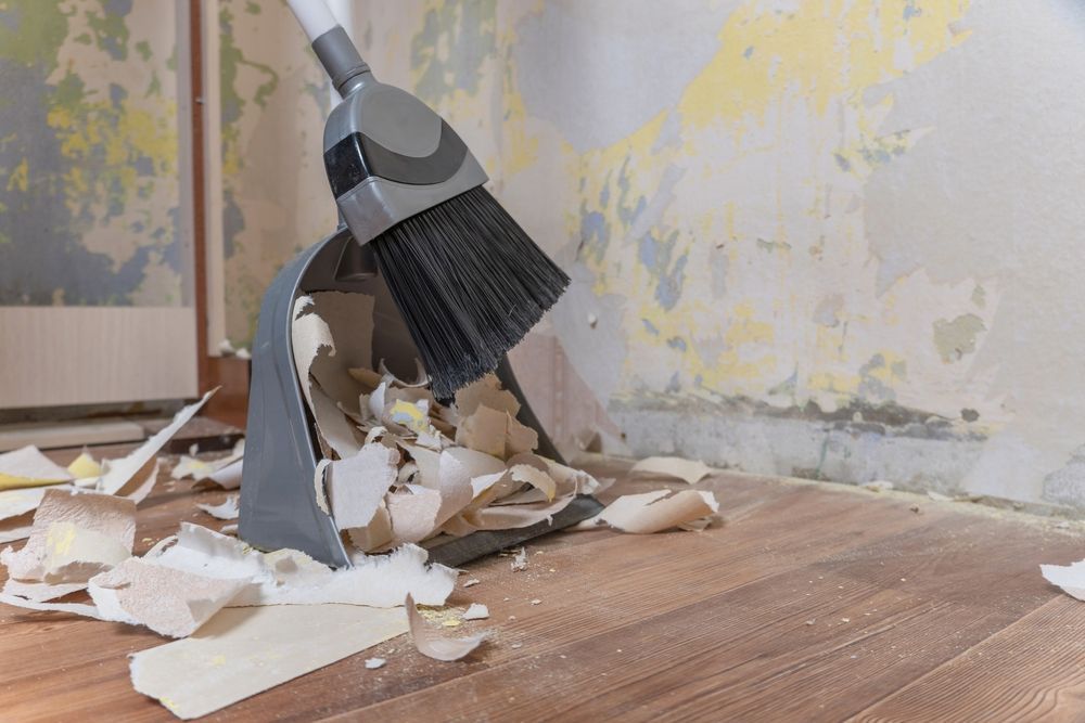 Broom sweeping debris into a dustpan on a wooden floor in a room with damaged walls.