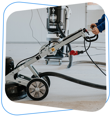 Person operating a floor grinding machine, with a vacuum cleaner in the background, in a construction setting.
