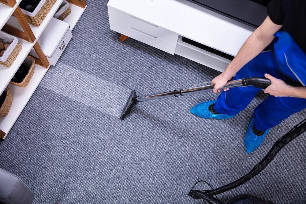 Person in blue coveralls cleans a gray carpet with a carpet cleaner.