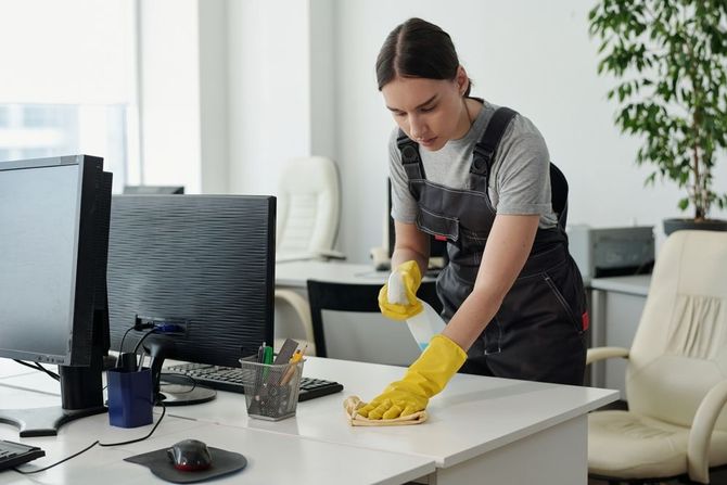 Woman in overalls and yellow gloves cleaning an office desk with spray and cloth.