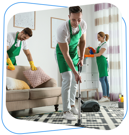 Three people in green aprons cleaning a living room. One vacuums, another dusts a couch, and the third carries cleaning supplies.