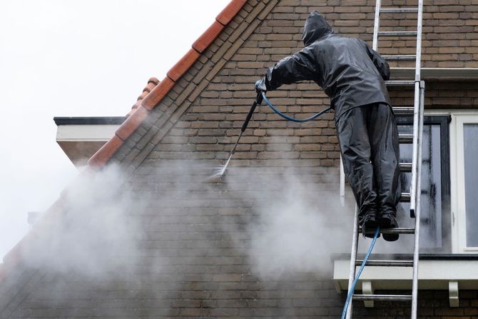 Person on ladder power washing brick house, spraying mist.