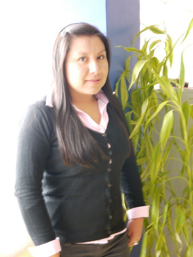 Una mujer con un suéter negro y una camisa rosa está parada frente a una planta.