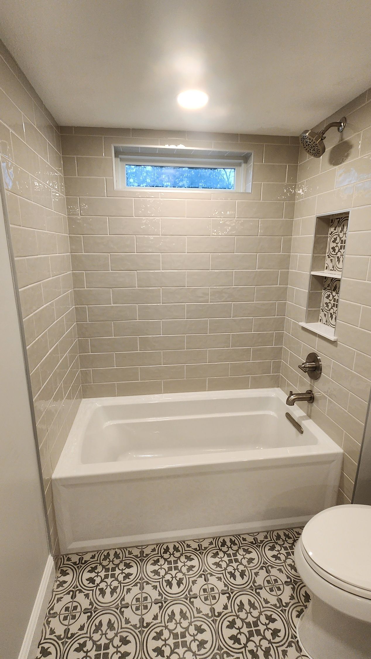 Small bathroom with white tub, beige tiled walls, patterned floor, and a small window.