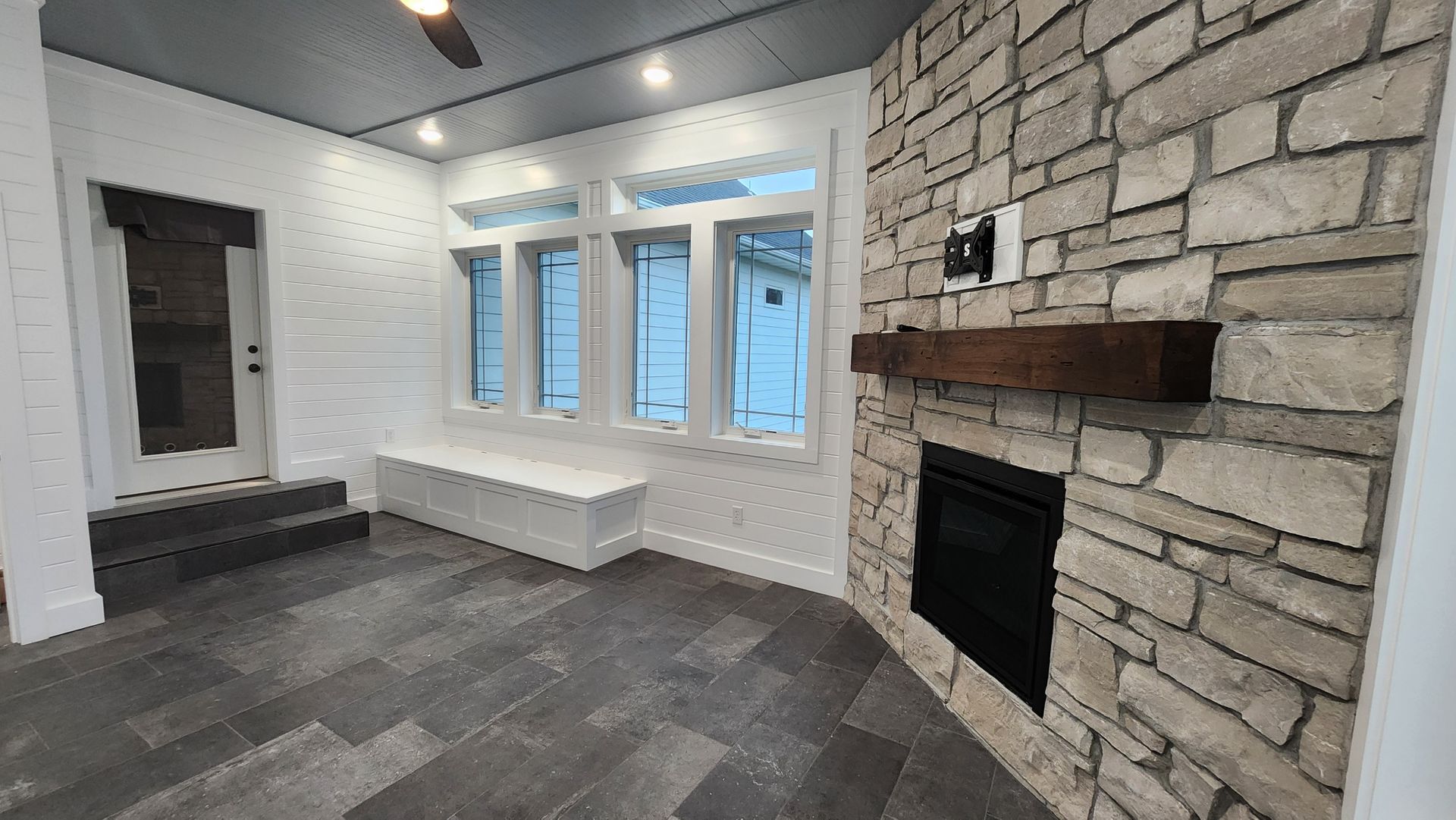 A room with gray stone fireplace, windows with white bench, and dark gray flooring.