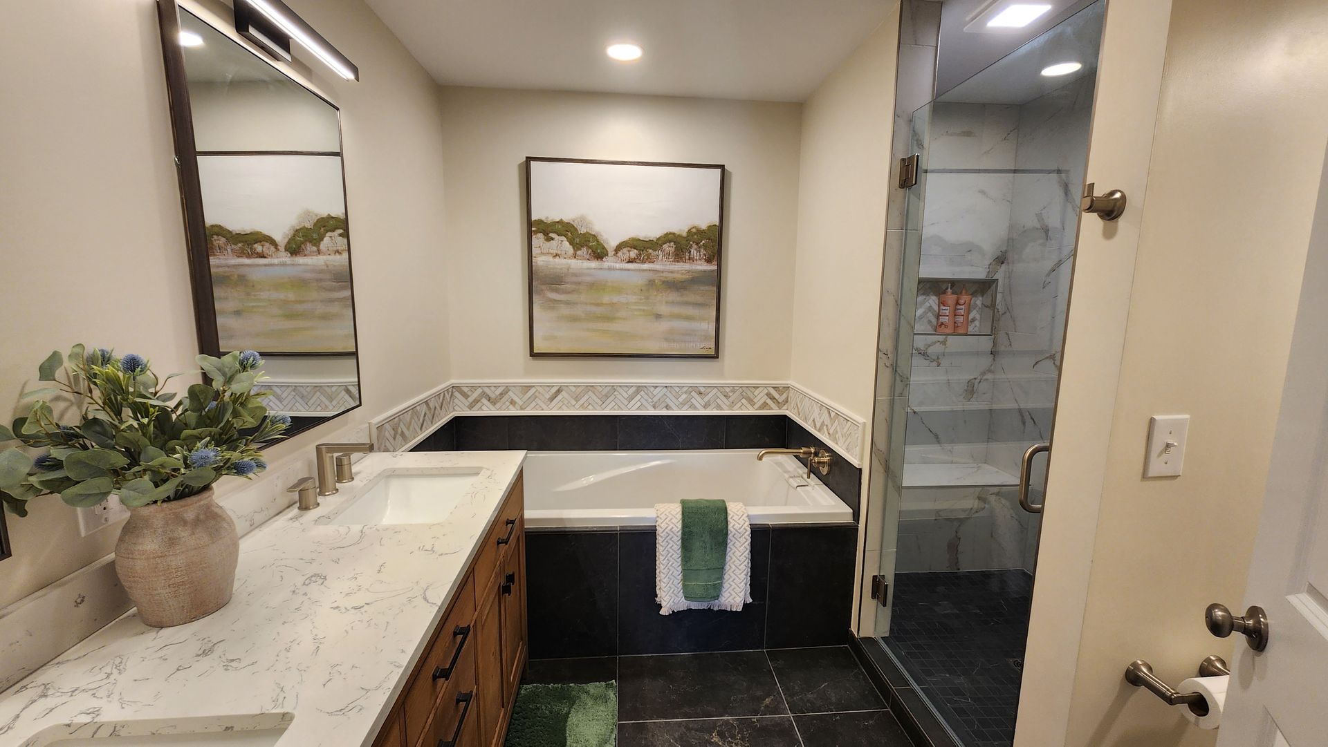 Bathroom with a bathtub, shower, vanity, and artwork. Light walls, dark tile, and stone countertops.