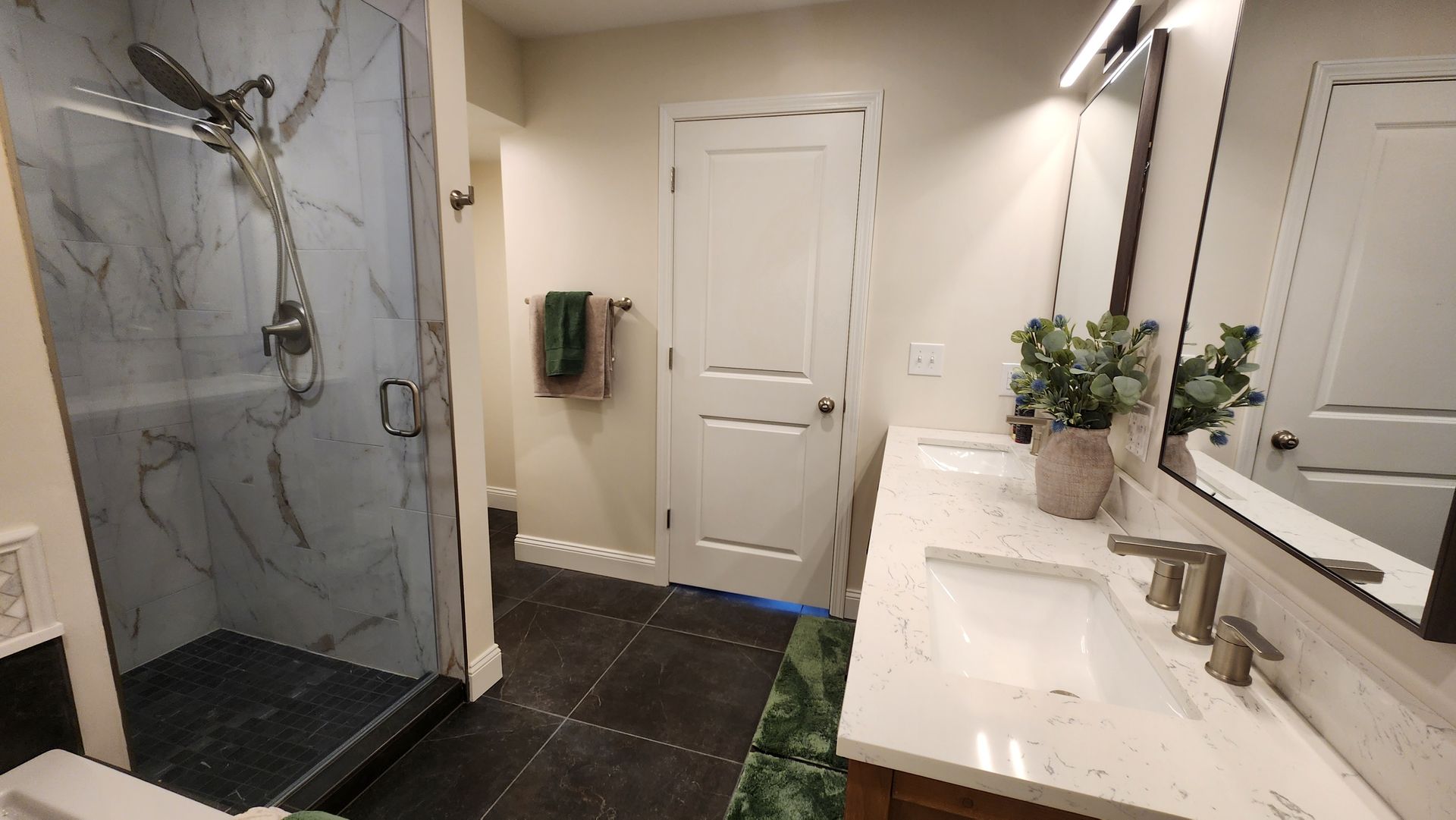 Bathroom with glass shower, marble tile, white vanity, dark floor, white door.
