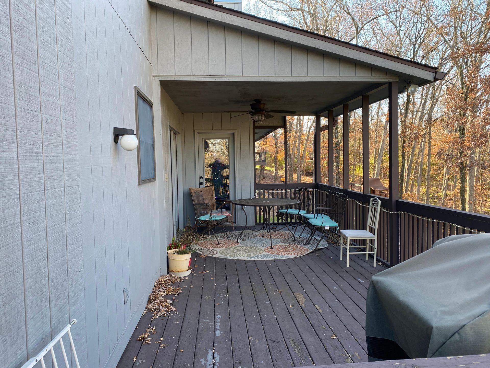 Wooden deck with a covered porch. A table and chairs sit on a rug. The house exterior is gray. Trees are in the background.