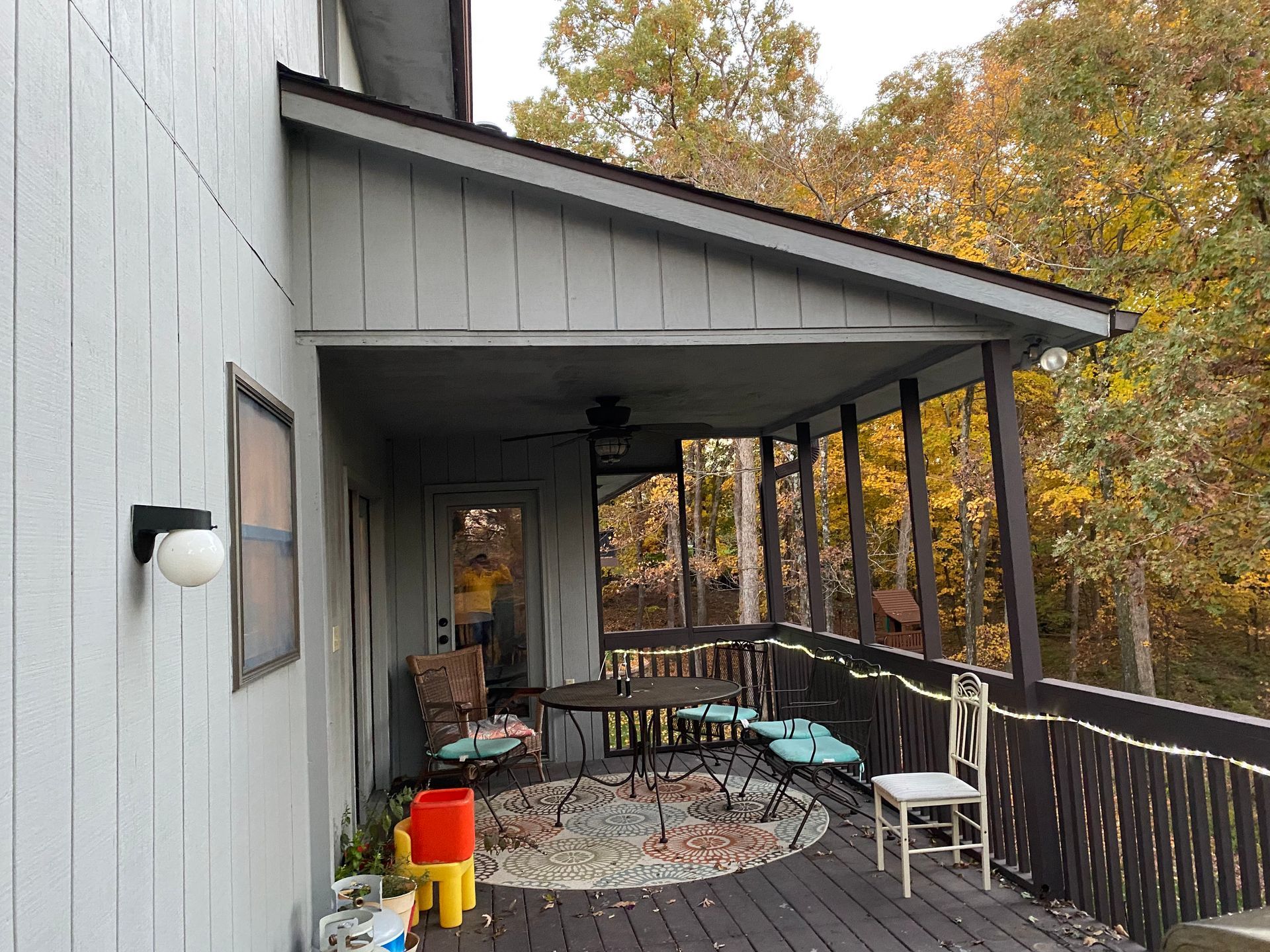 Covered porch with seating; a gray house with a dark brown railing and an autumnal forest in the background.