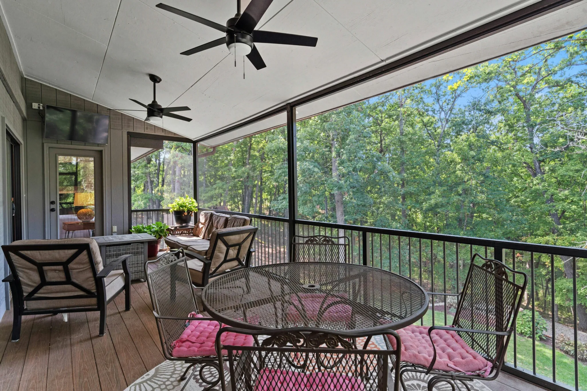 Screened-in porch with outdoor furniture, overlooking a wooded area. Ceiling fans and decorative iron table.