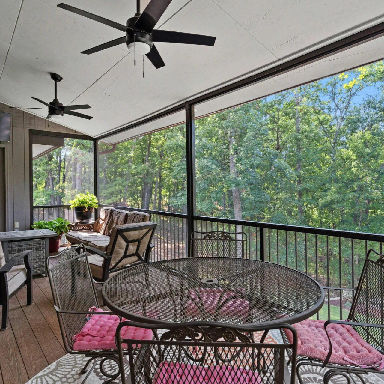 Screened-in porch with metal furniture, pink cushions, and forest view. Two ceiling fans.