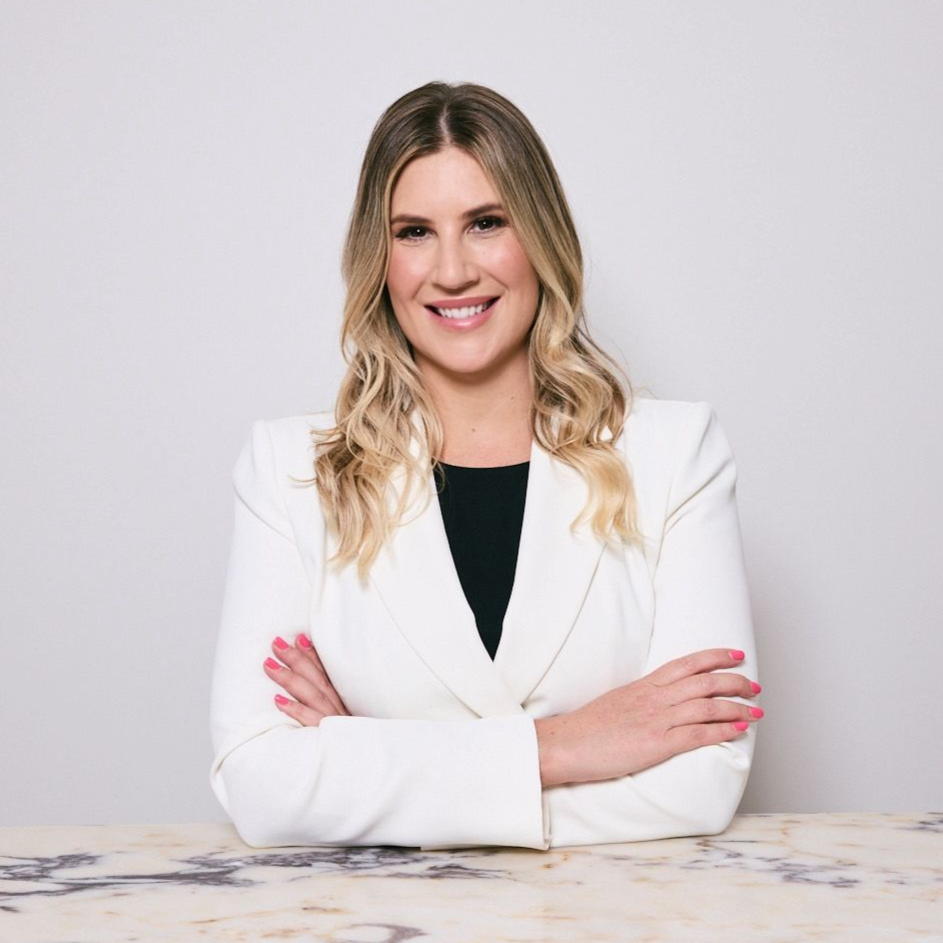 Woman with blonde hair, wearing a white blazer and black top, smiling with arms crossed.