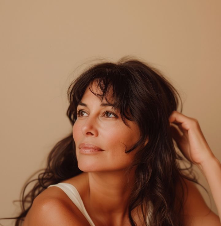Woman with dark wavy hair, looking away thoughtfully, hand in hair, soft lighting, neutral background.