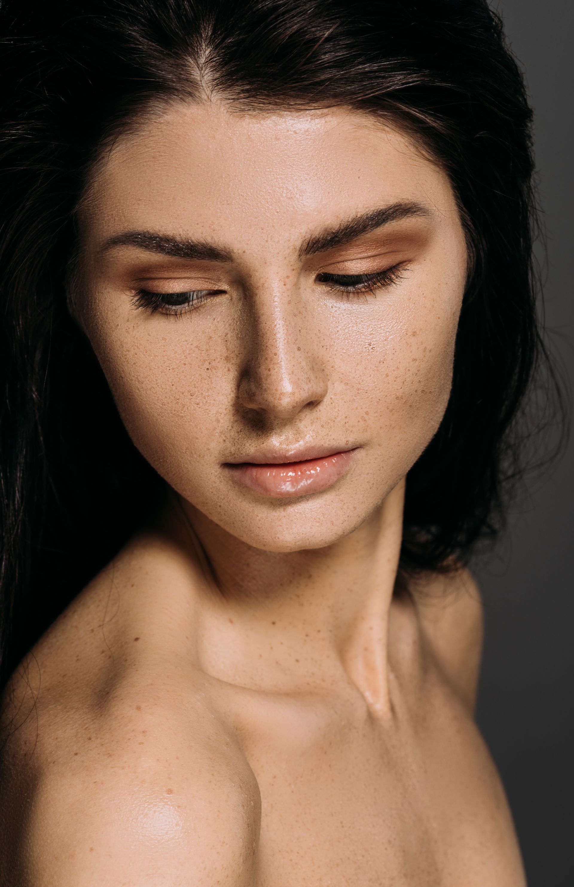 Woman with dark hair and freckles, looking down with a neutral expression. Brown eyeshadow, bare shoulders.