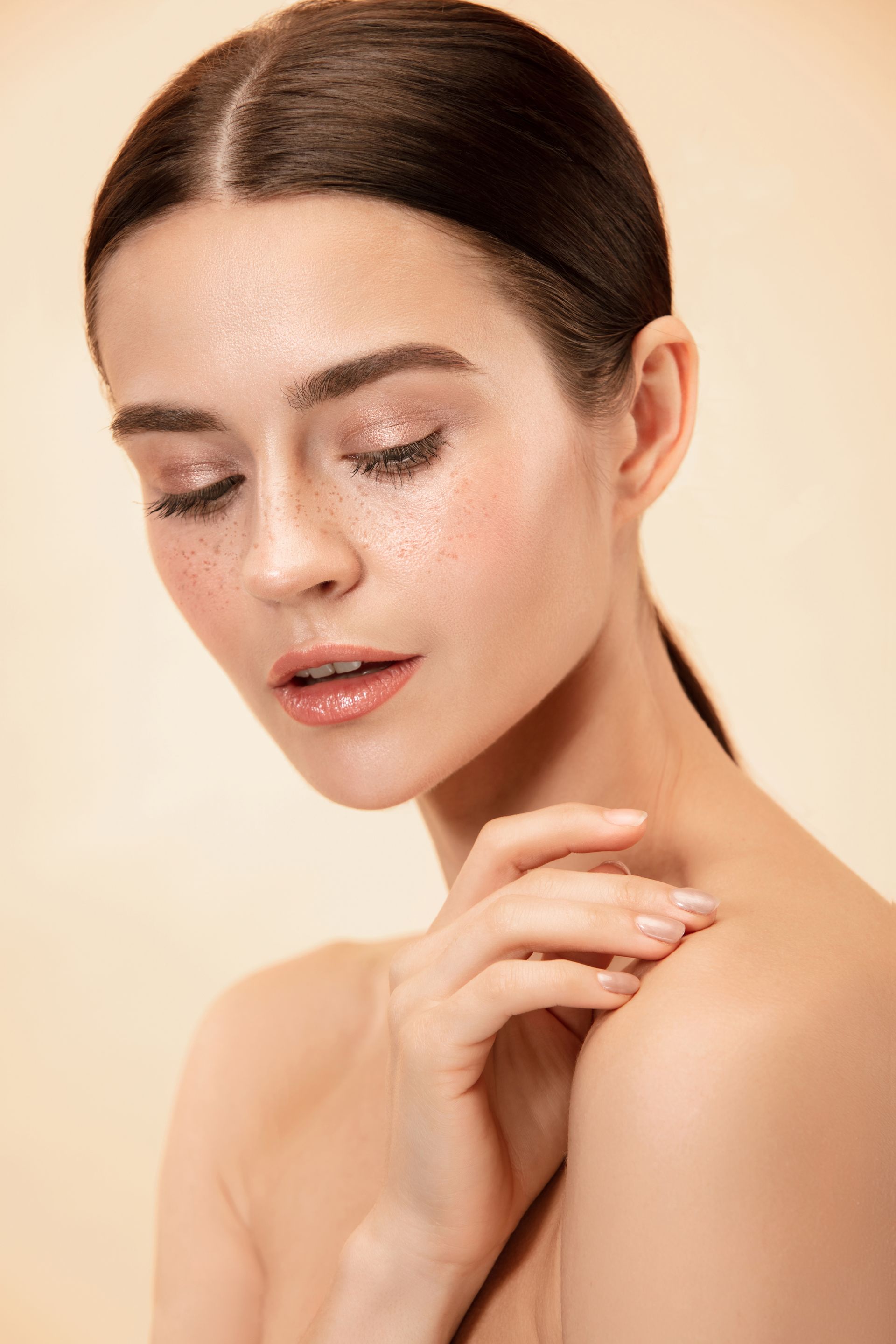 Woman with slicked-back hair, eyes closed, hand on shoulder, glowing skin, neutral background.