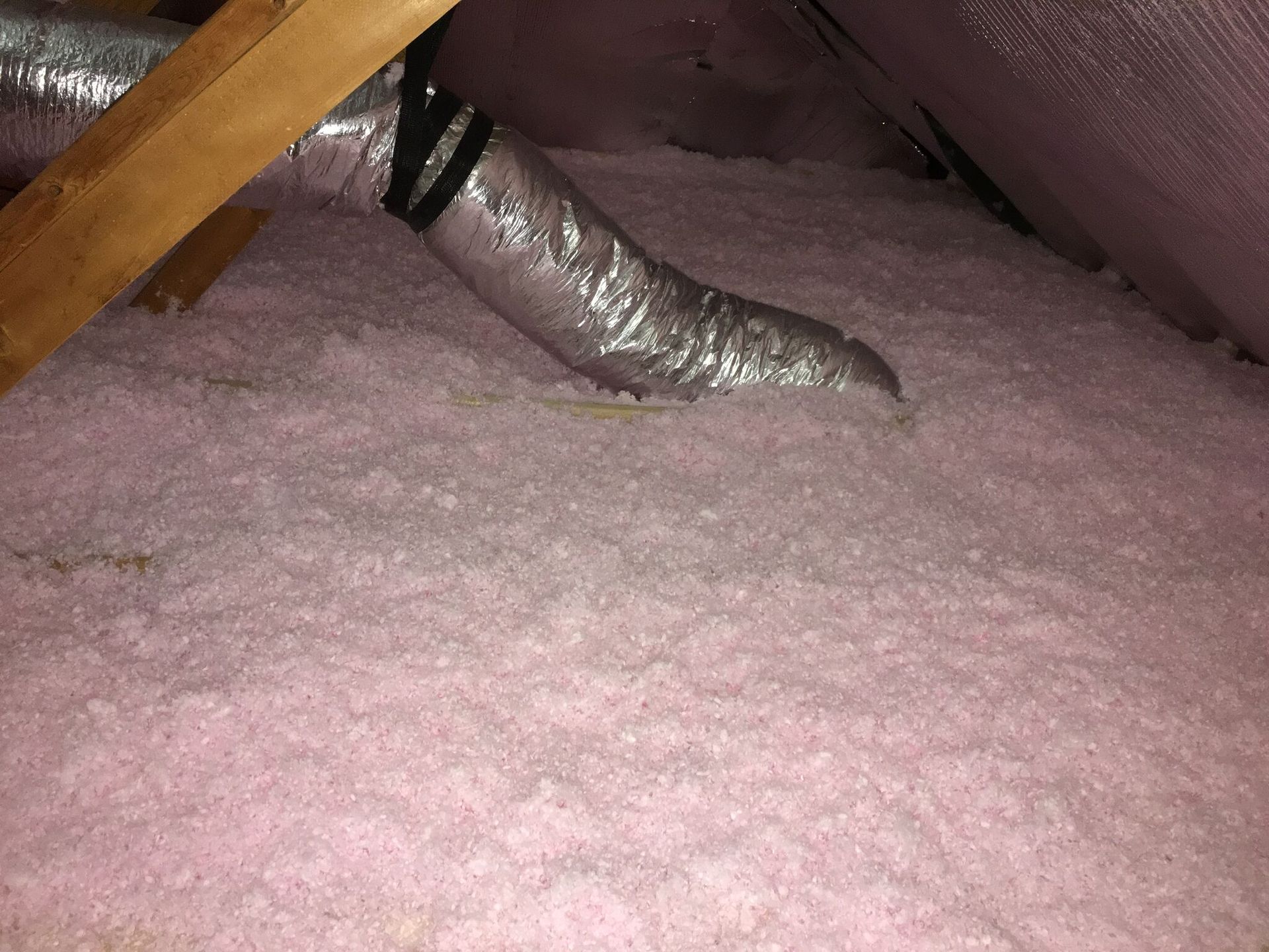 Pink insulation fills an attic, with a silver HVAC duct and wood beams visible.