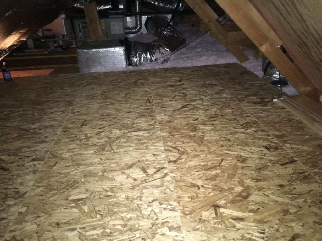 Plywood flooring in an attic with HVAC equipment in the background and angled wooden beams.
