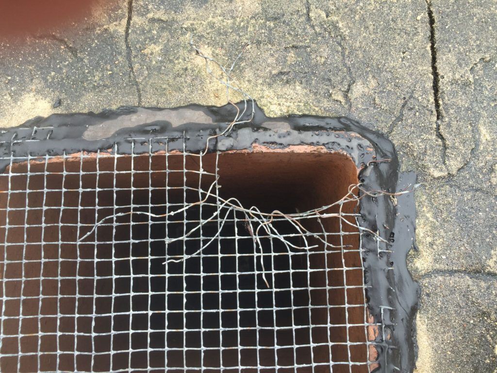 Close-up of a damaged wire mesh cover over a chimney opening, attached to a roof with sealant.