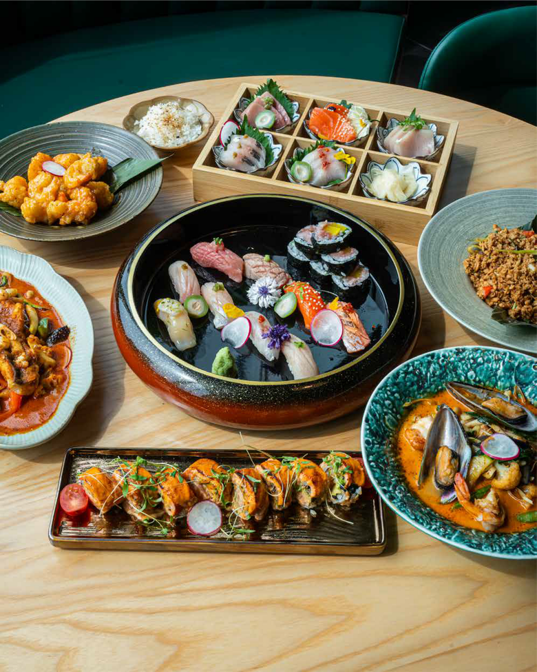 A wooden table spread with a variety of Asian dishes, including sashimi, sushi rolls, seafood bowls, and fried rice.