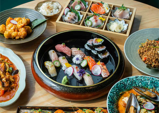 A wooden table spread with a Japanese meal including sushi, sashimi, fried shrimp, a rice bowl, and seafood in curry sauce.