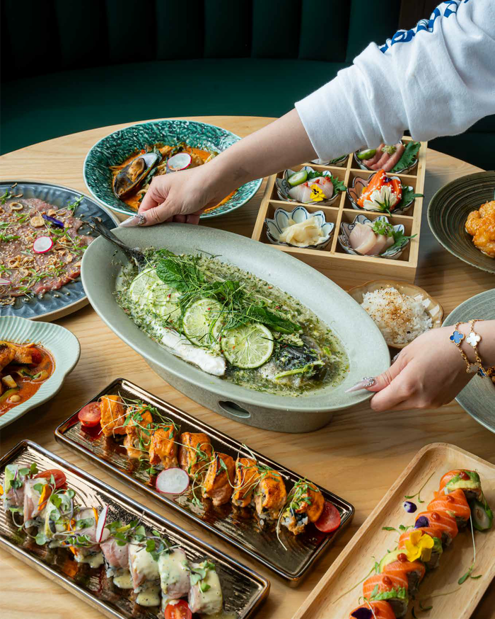 Hands serve a spread of various seafood dishes, including sushi, sashimi, and a whole fish in lime sauce, on a table.