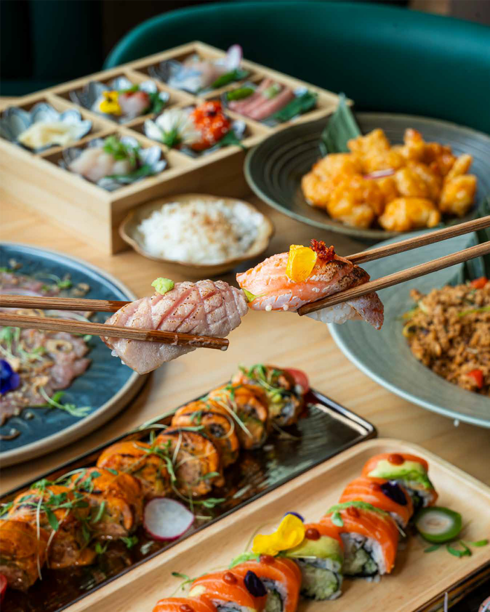 Chopsticks hold two pieces of sushi over a table filled with assorted sushi, fried food, and a bowl of rice.