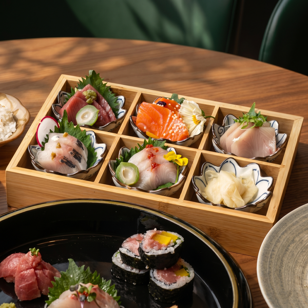 A wooden serving box containing six varieties of fresh sashimi and ginger, next to a black plate with sushi rolls.