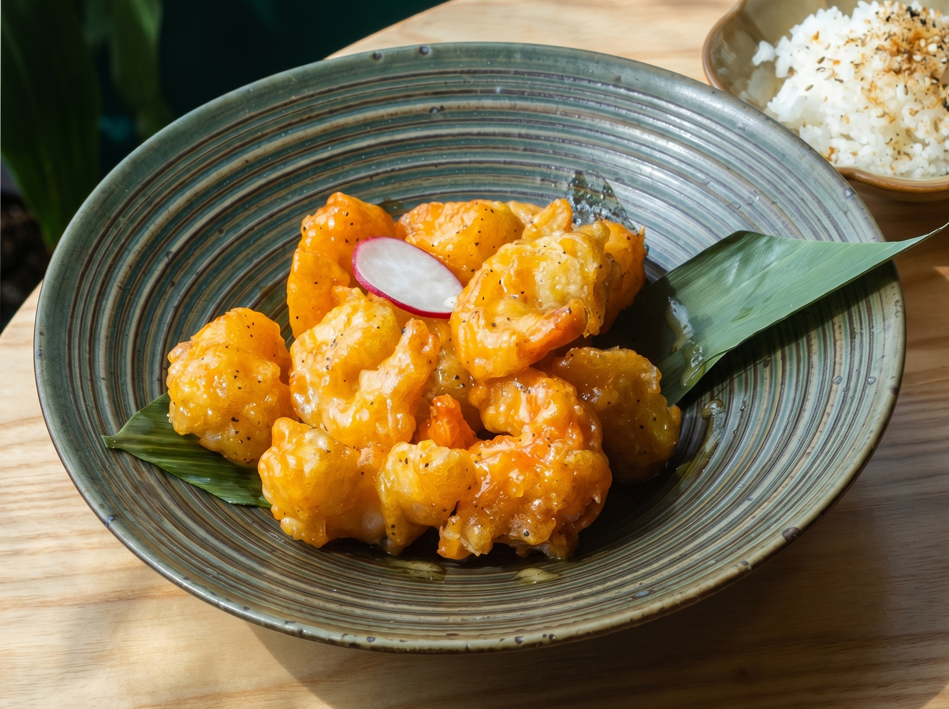 A bowl of golden, sauce-glazed fried shrimp garnished with a radish slice and a green leaf, served with side rice.