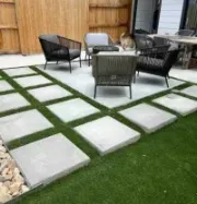 Modern backyard patio with gray paver stones, artificial grass, and outdoor furniture, bordered by a wooden fence.