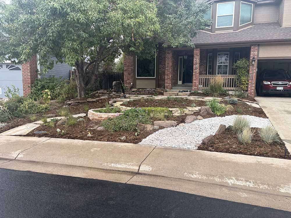 A suburban house with a landscaped front yard featuring mulch, a gravel pathway, stone edging, and a large tree.