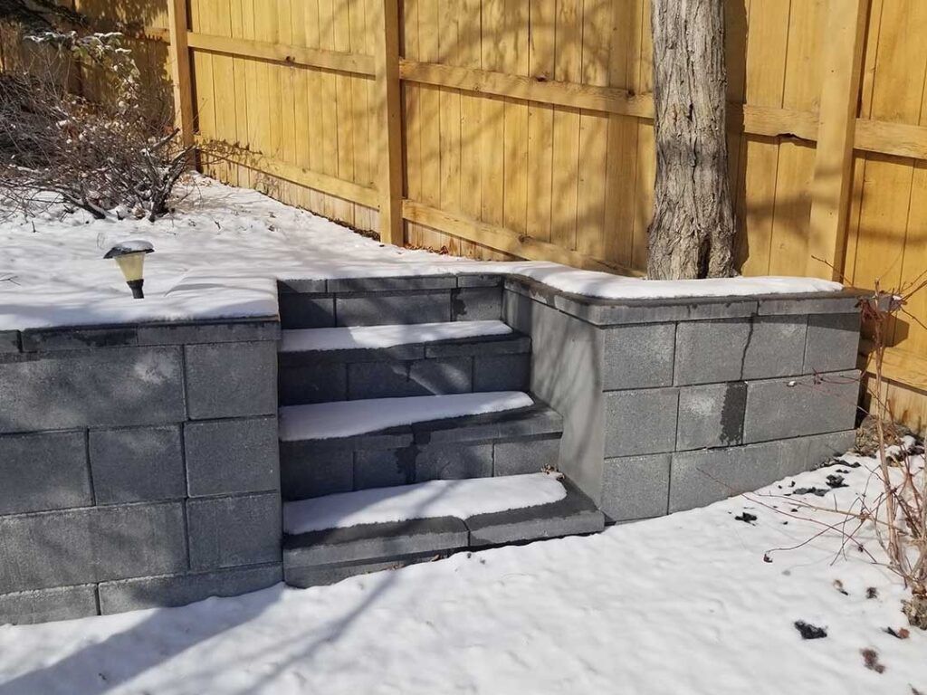 A set of stone steps built into a retaining wall in a snow-covered backyard.
