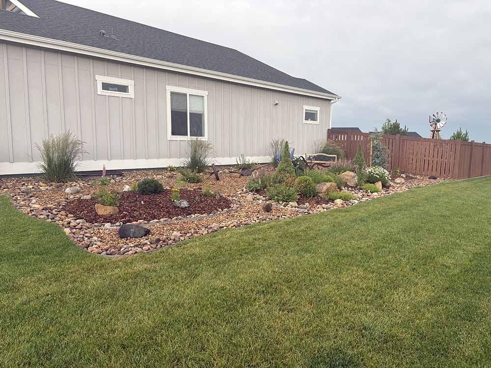 A light-grey house with a rocky landscape bed in the front yard and a brown fence along the side.