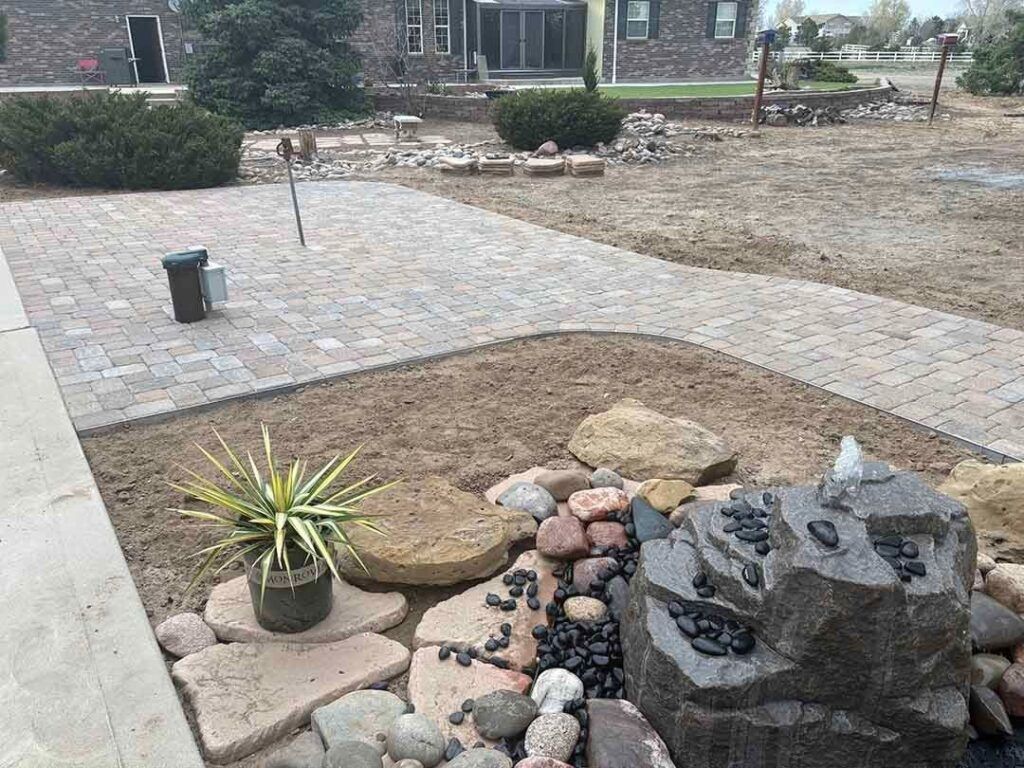 A paved patio leads toward a house, with a stone water feature, decorative rocks, and a small potted plant in the foreground.