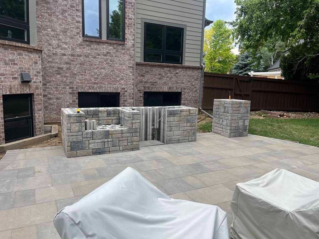 A stone outdoor kitchen and island being built on a light-colored paver patio near a brick house, with two covered chairs.