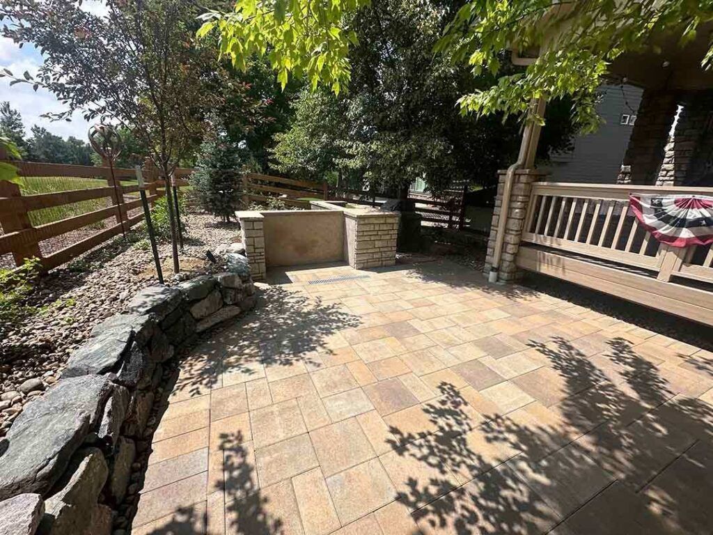 A stone patio with a built-in outdoor kitchen, stone retaining wall, and a wooden deck under trees in a backyard.