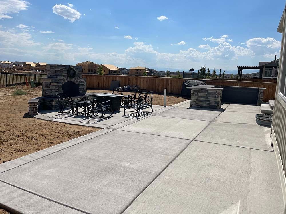 A backyard patio with a stone fireplace, outdoor furniture, and a stone kitchen counter under a bright blue sky.