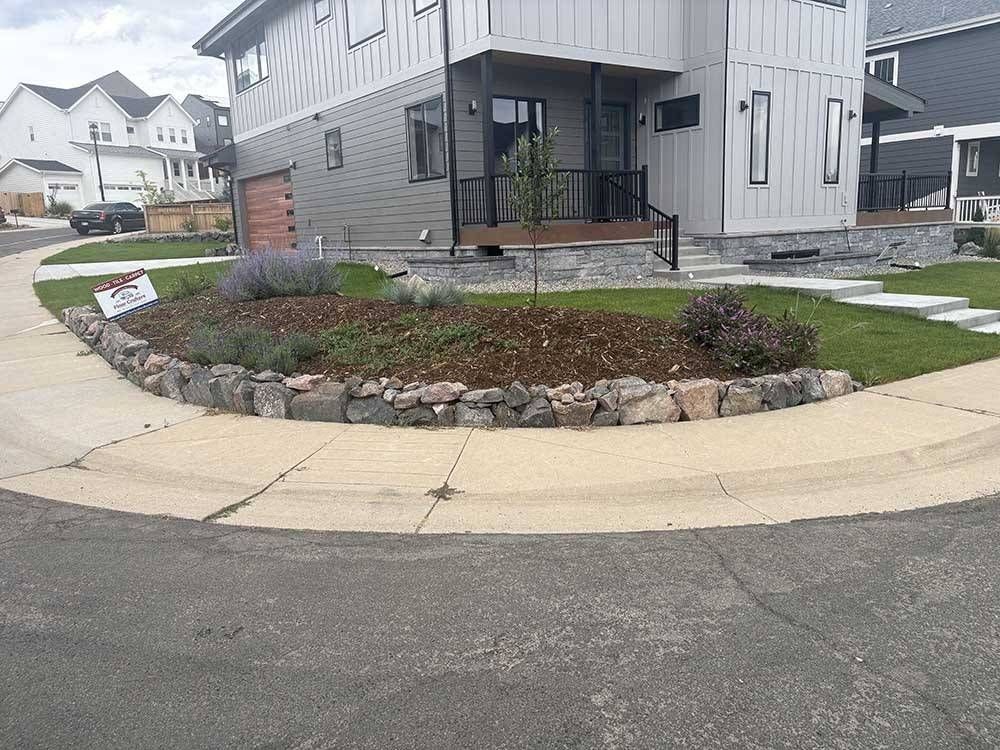 A residential corner lot with a curved rock garden bed featuring small shrubs, mulch, and a grey house in the background.