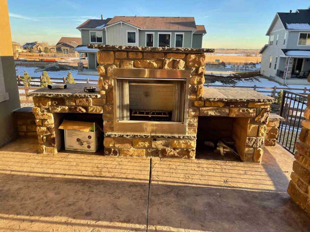 A tan stone outdoor fireplace with two built-in storage alcoves, situated on a patio overlooking a suburban neighborhood.