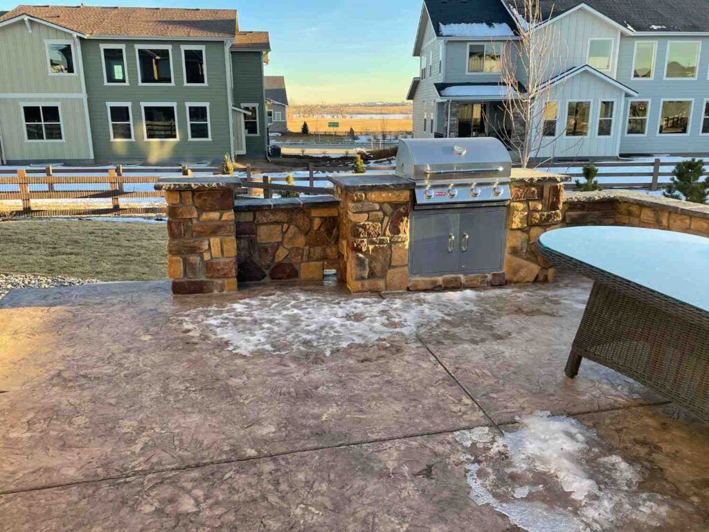 A backyard patio featuring a stone-clad outdoor kitchen with a built-in grill, next to a table and houses.