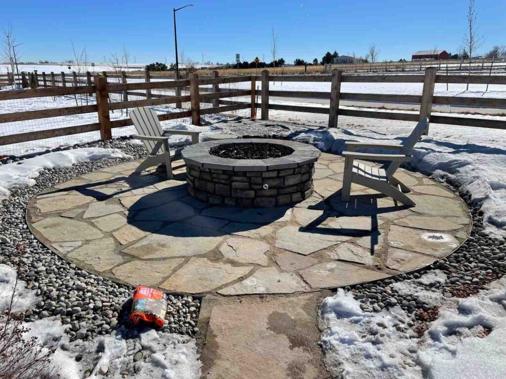 A stone fire pit with two chairs on a circular flagstone patio, surrounded by snow and a wooden fence on a sunny day.