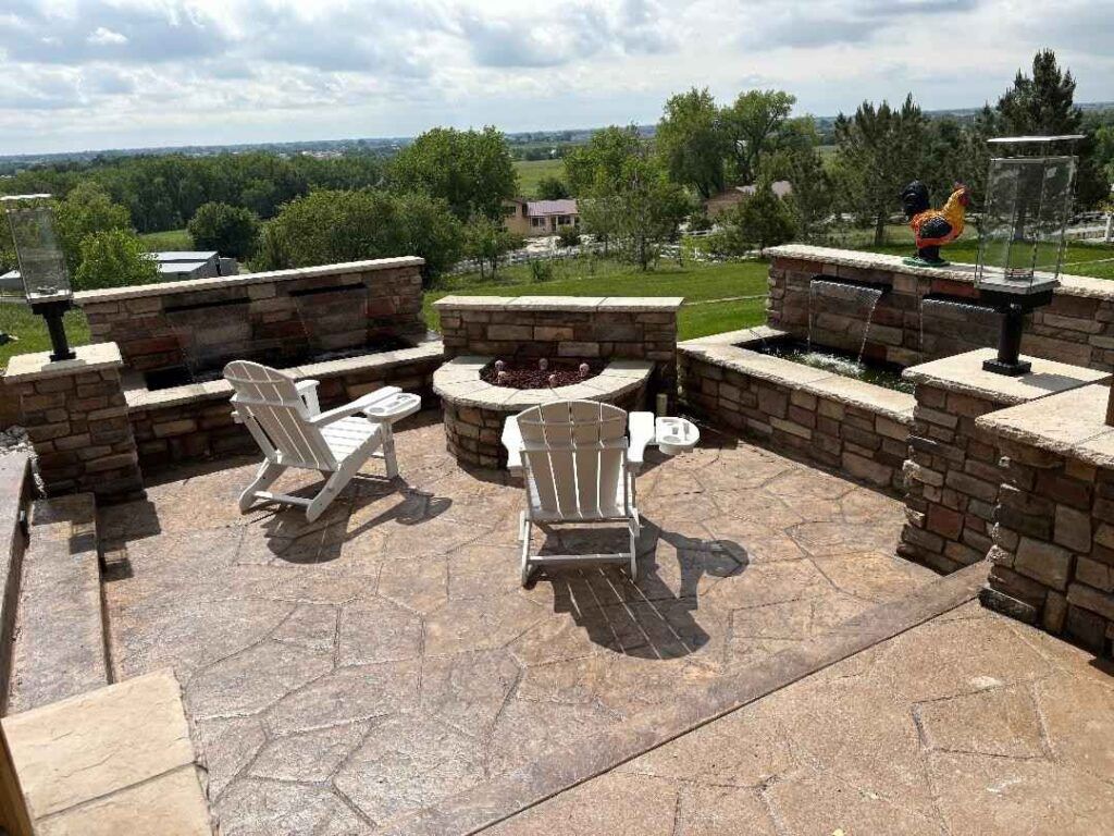 A stone patio with a central fire pit, two white chairs, and two water features, overlooking a green, rural landscape.