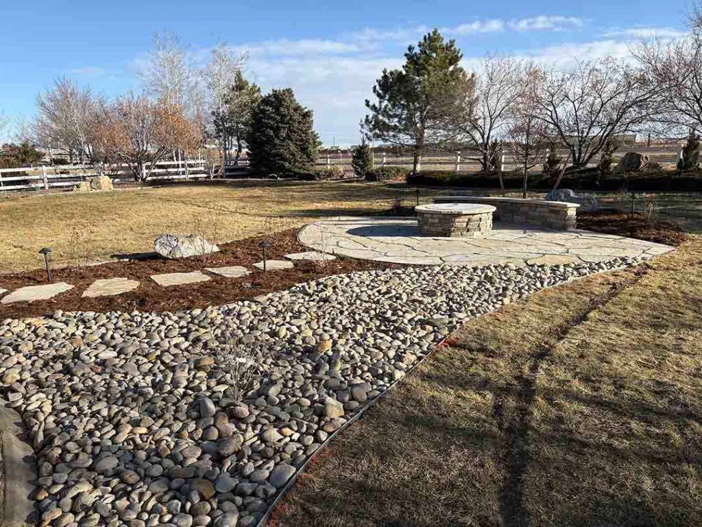 A stone patio with a fire pit, adjacent to a rock-filled drainage bed and stepping stones in a grassy backyard.