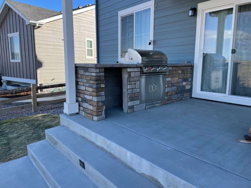 A stone-clad outdoor kitchen with a built-in stainless steel grill on a concrete patio beside a modern blue-sided house.