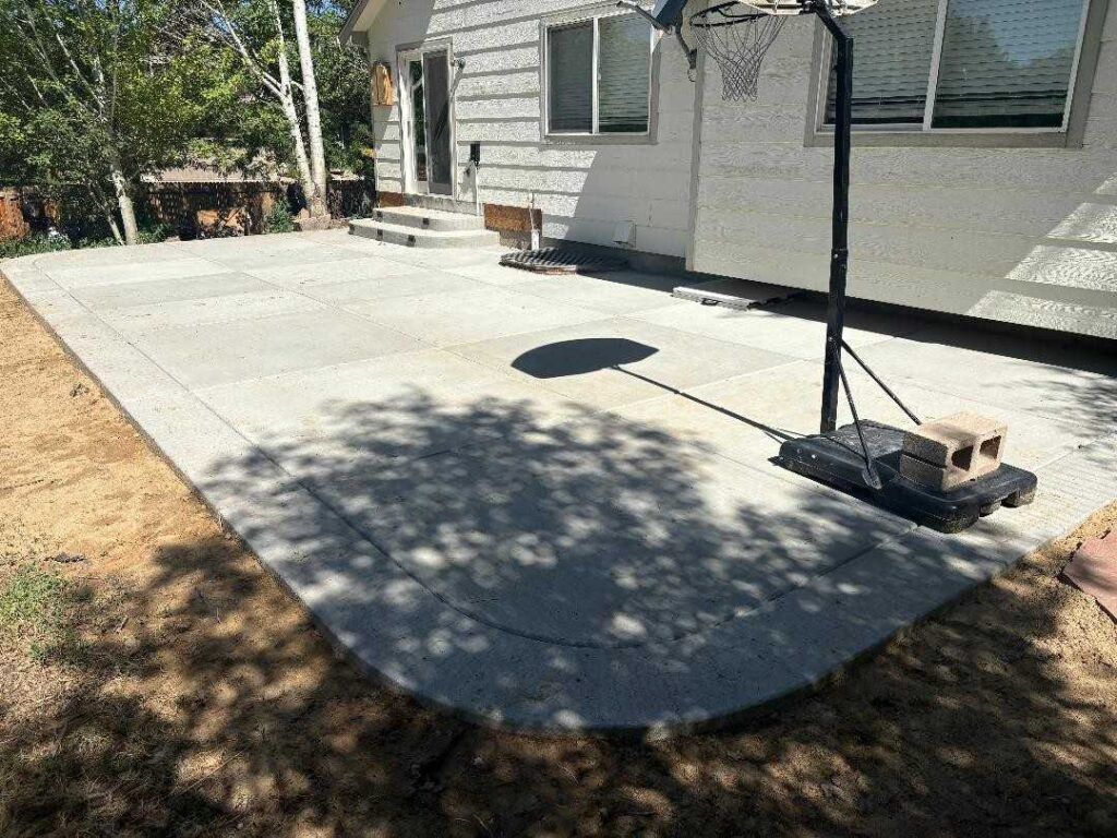 A light-colored concrete patio adjacent to a house exterior, featuring a portable basketball hoop on the right side.