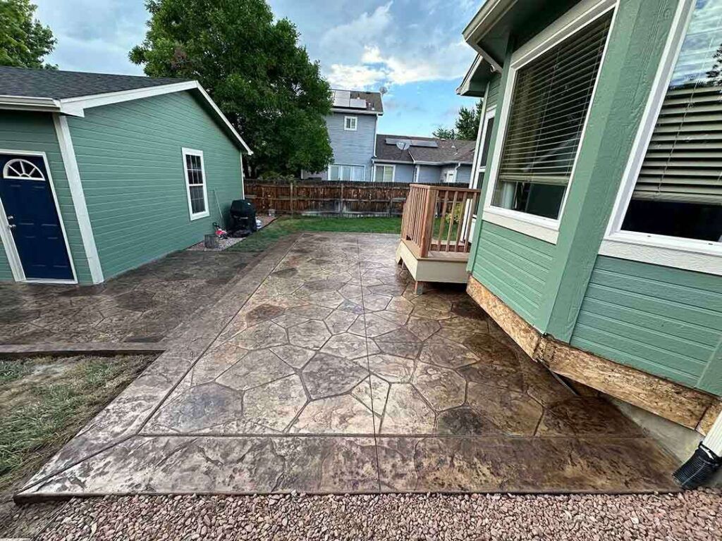 A stamped concrete patio featuring a flagstone pattern with a border, adjacent to a house with sage-green siding.
