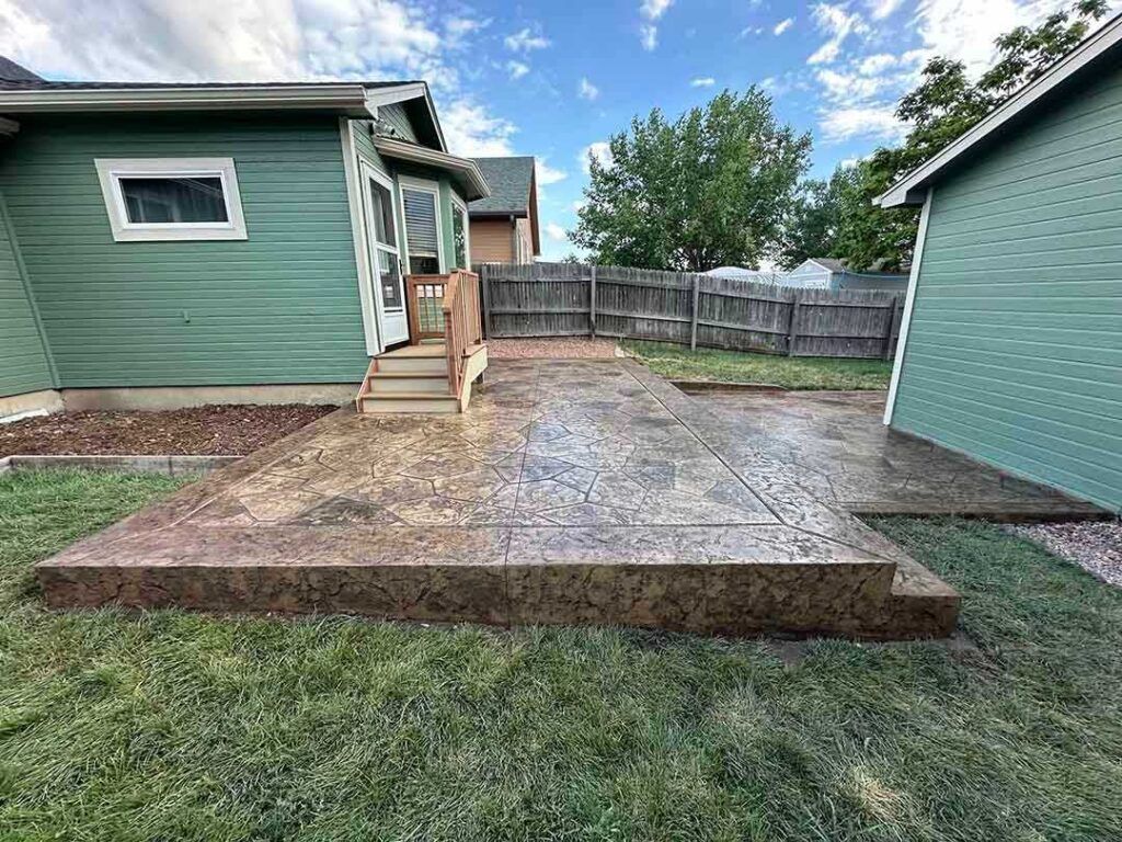 A brown stamped concrete patio outside a green house with a small wooden staircase leading to a door.